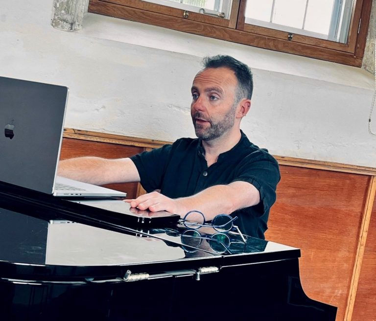 Jamie Read Singing Lessons and Vocal Coaching Jamie is looking at the screen of an Apple MacBook, which is on the top of a piano. He is teaching someone online