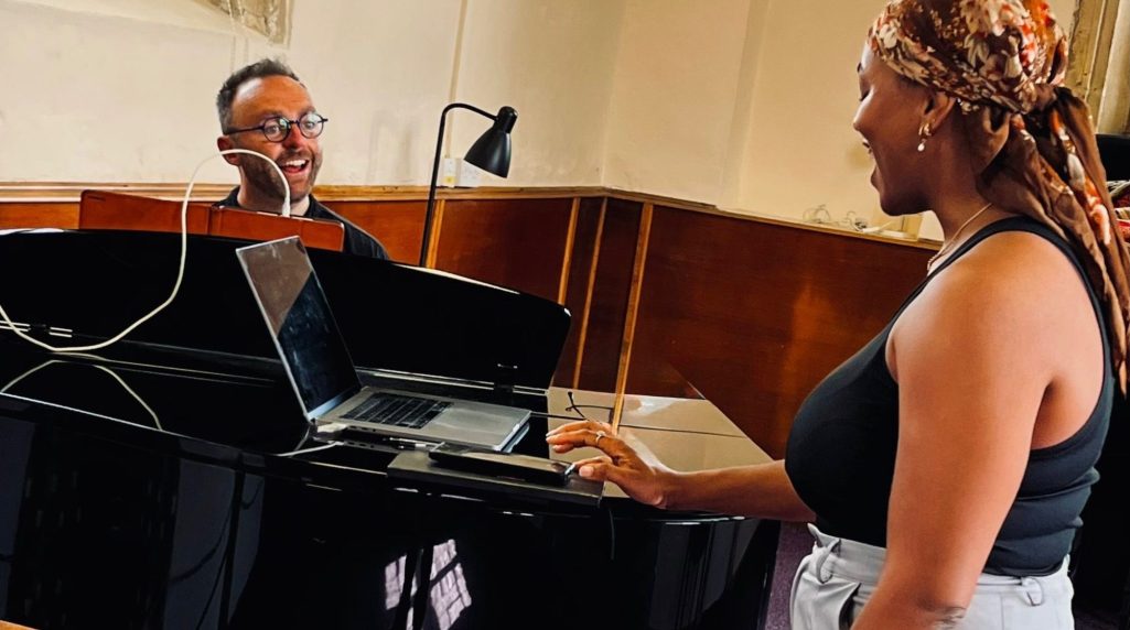 West End Vocal Coach, Jamie Read Jamie Read - a white, middle aged man with a beard - is sat at a grand piano smiling at a singer, who is a black woman in her mid thirties