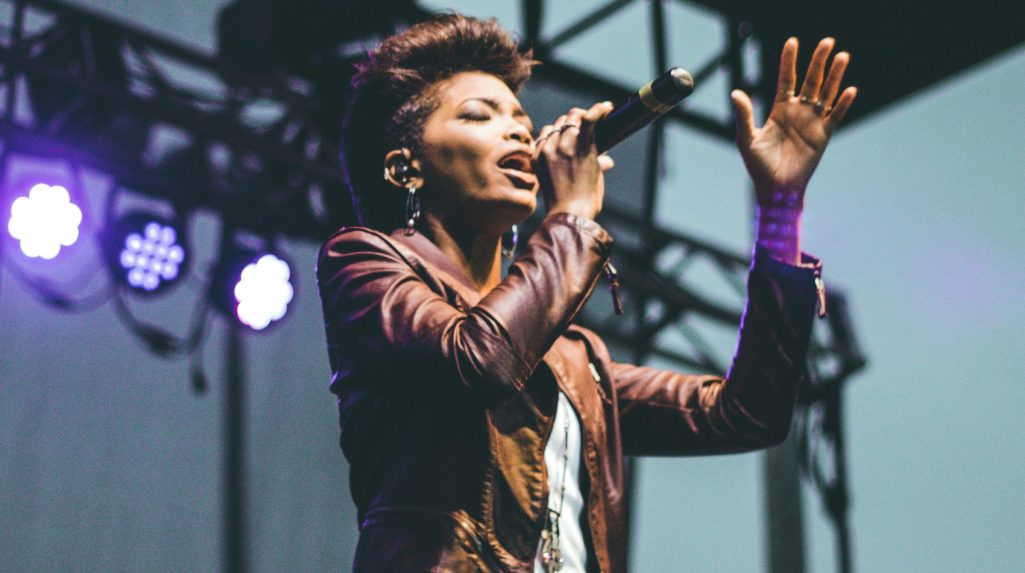 A photo showing a singer on stage. The singer is a black woman holding a mic, with stage lights in the background