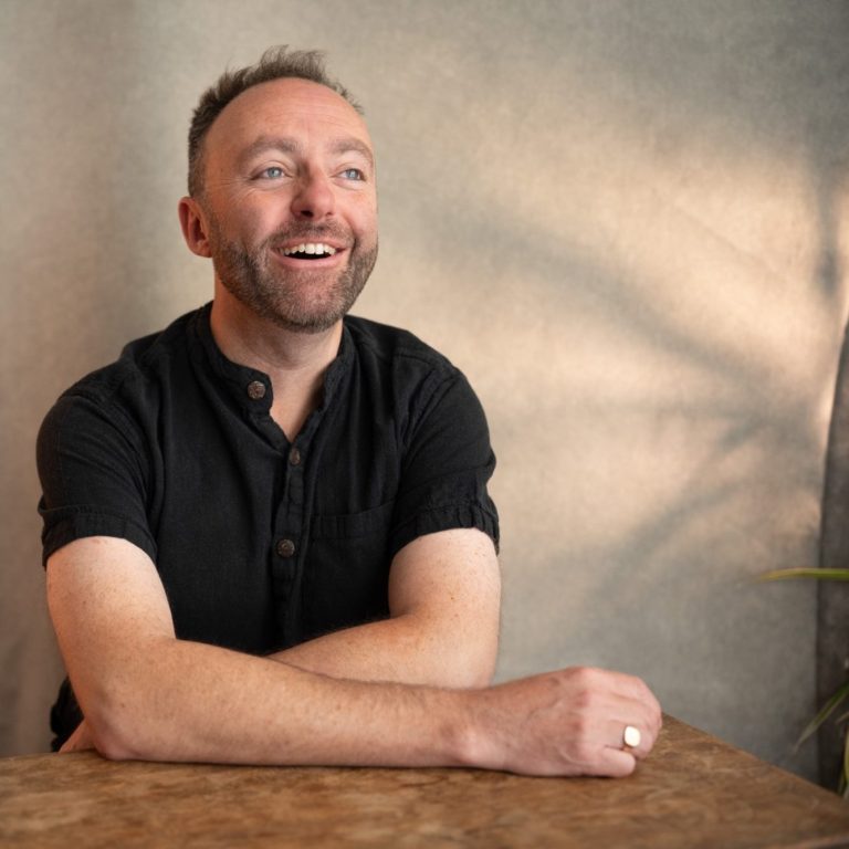 Jamie Read is a Vocal Coaching and Singing Teacher in Thatcham A photo of the vocal coach, Jamie Read, laughing towards the camera. He is wearing a black shirt and leaning on a wooden table